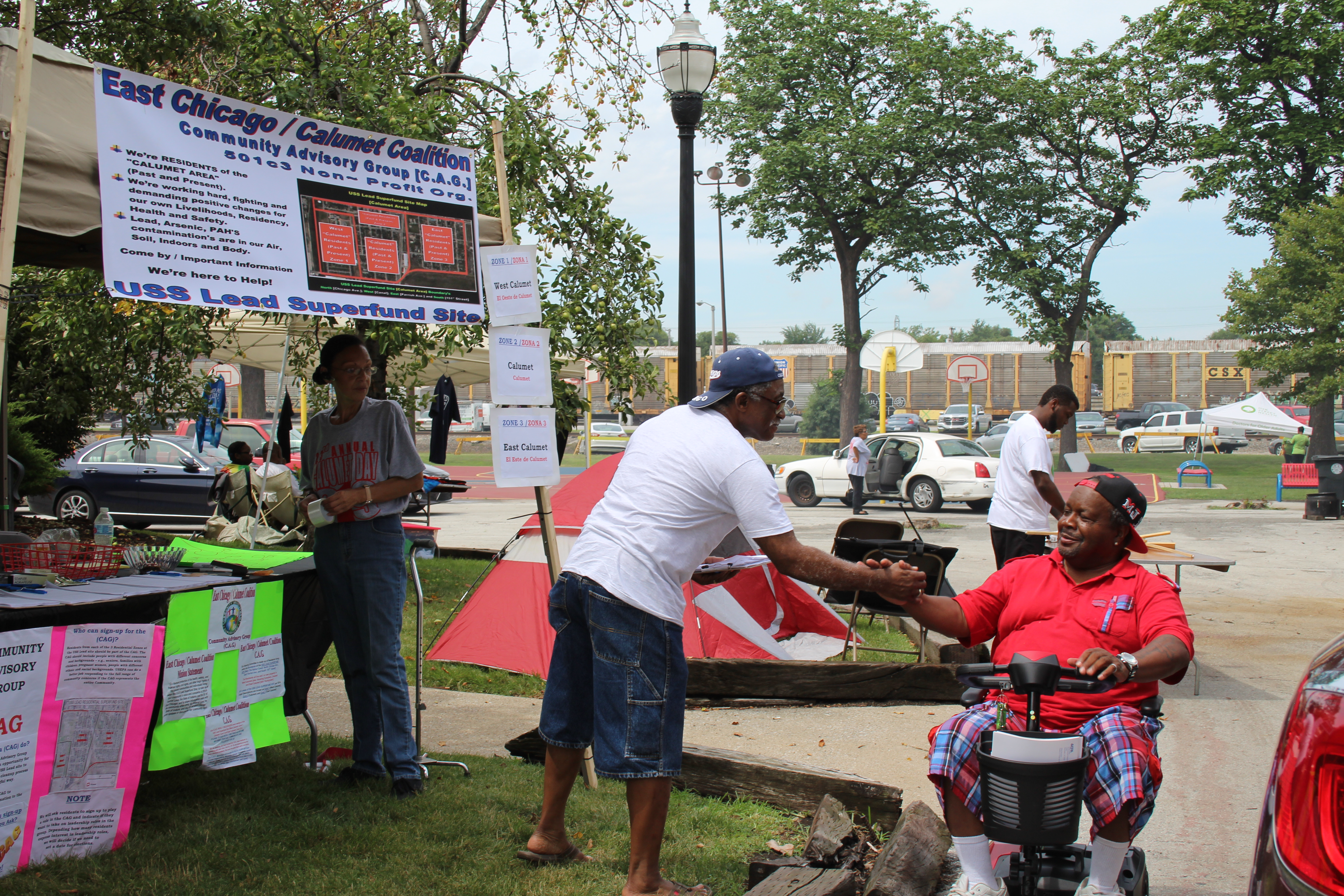 The East Chicago Community Advisory Group has a stand at an annual park event. Two Black men in the foreground embrace one another.