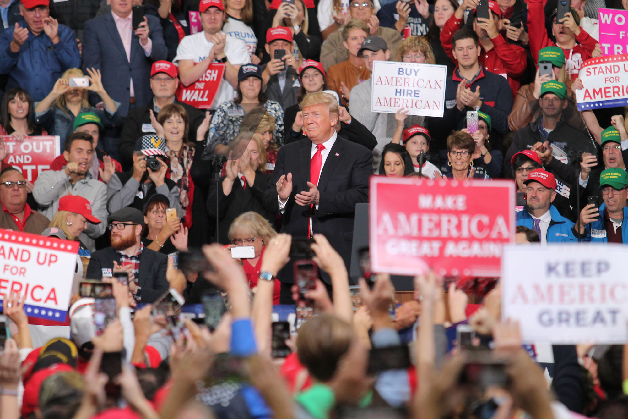 President Donald Trump claps as a crowd in front and behind him wave signs and cheer for him.