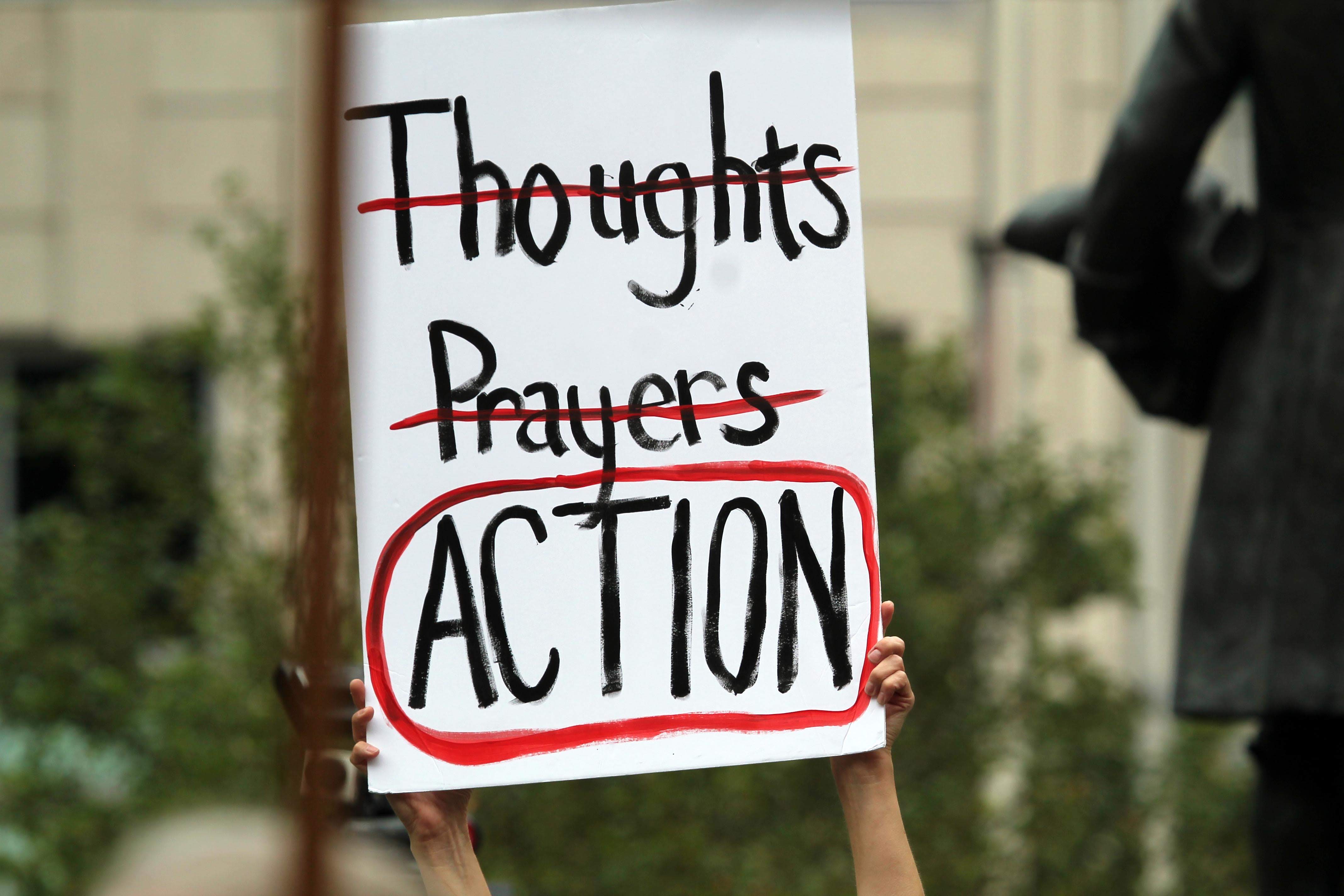 A person holds up a sign that reads 'Thoughts, Prayers, Actions' with thoughts and prayers struck through with red marker and action circled in the same color.