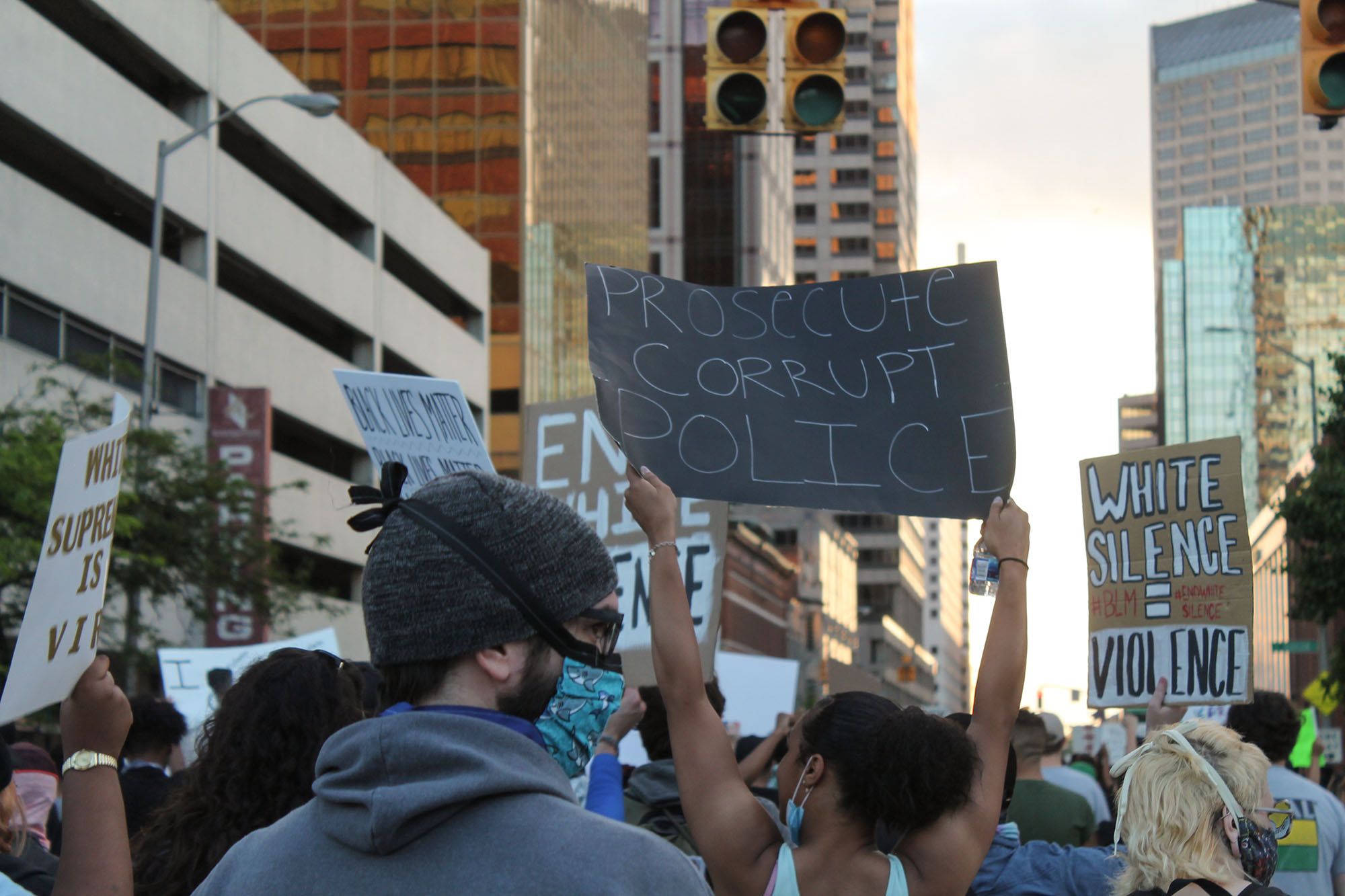 An immense crowd marches through downtown Indianapolis. Several signs are held in the air, but 'Prosecute corrupt police' is clearly visible and in focus.