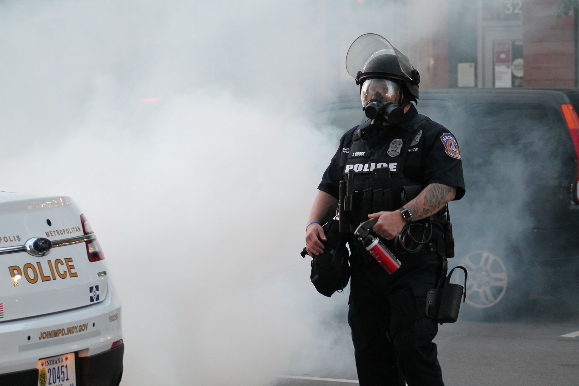 A police officer stands in front of an opaque cloud of tear gas. The officer wears riot gear and has a pepper spray cannister in their hand.