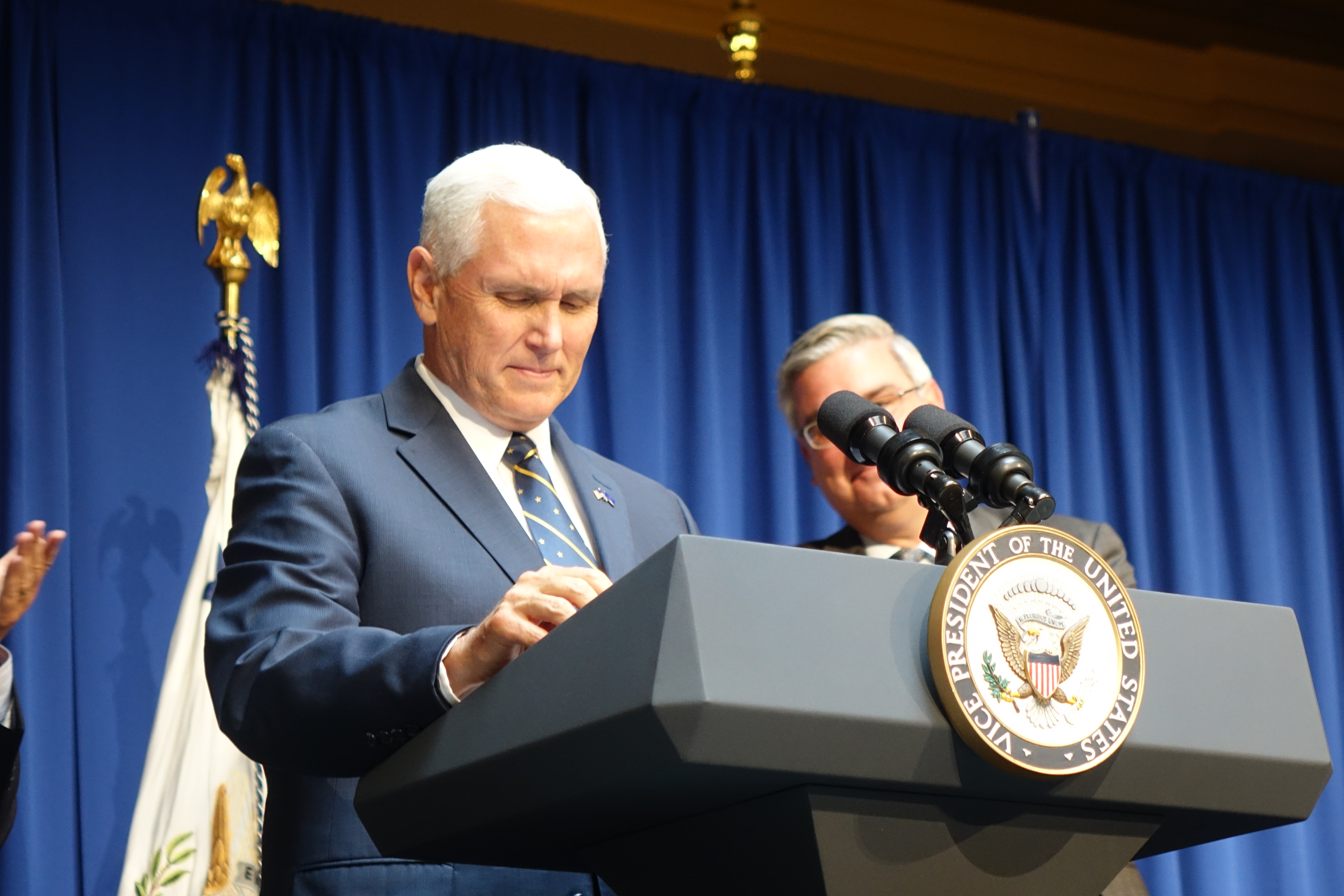 Mike Pence looks down at a podium with the vice presidential seal on it. Behind him, partially obscured by the mircrophone, is former Governor Eric Holcomb, his former lieutenant governor.
