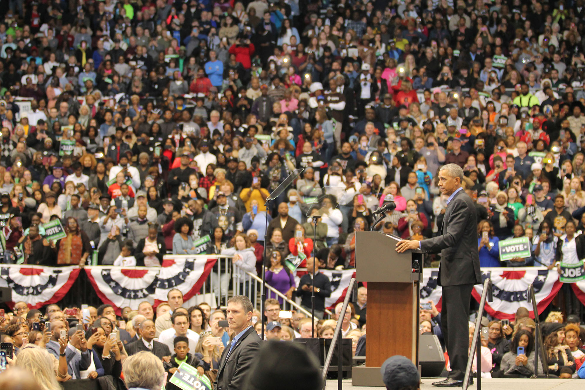 Barack Obama stands in front of a podium. Behind him are hundreds of people in a crowd, listening to his speech.
